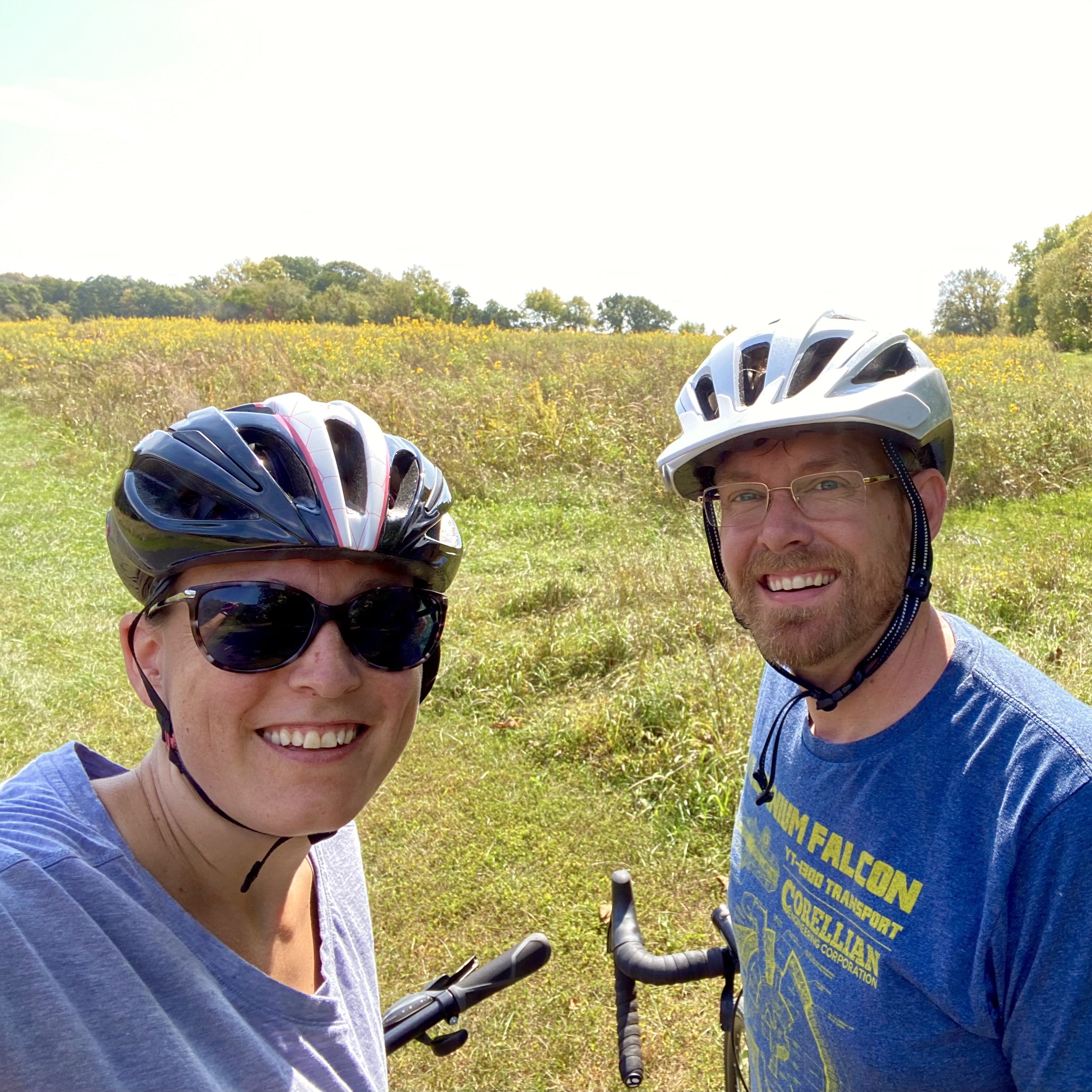 Courtney Tripp is pictured wearing a bike helmet and sunglasses. She is smiling and standing next to her husband Asa who is also wearing his bike helmet while biking on a Kansas trail.